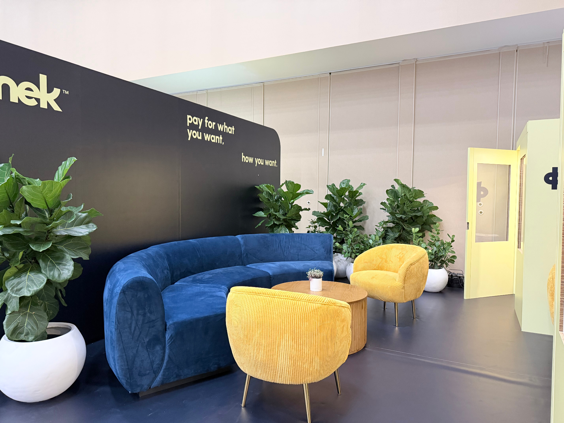 Konek private lounge with black backdrop, blue velvet sofa, yellow corduroy chairs, and fiddle-leaf planting